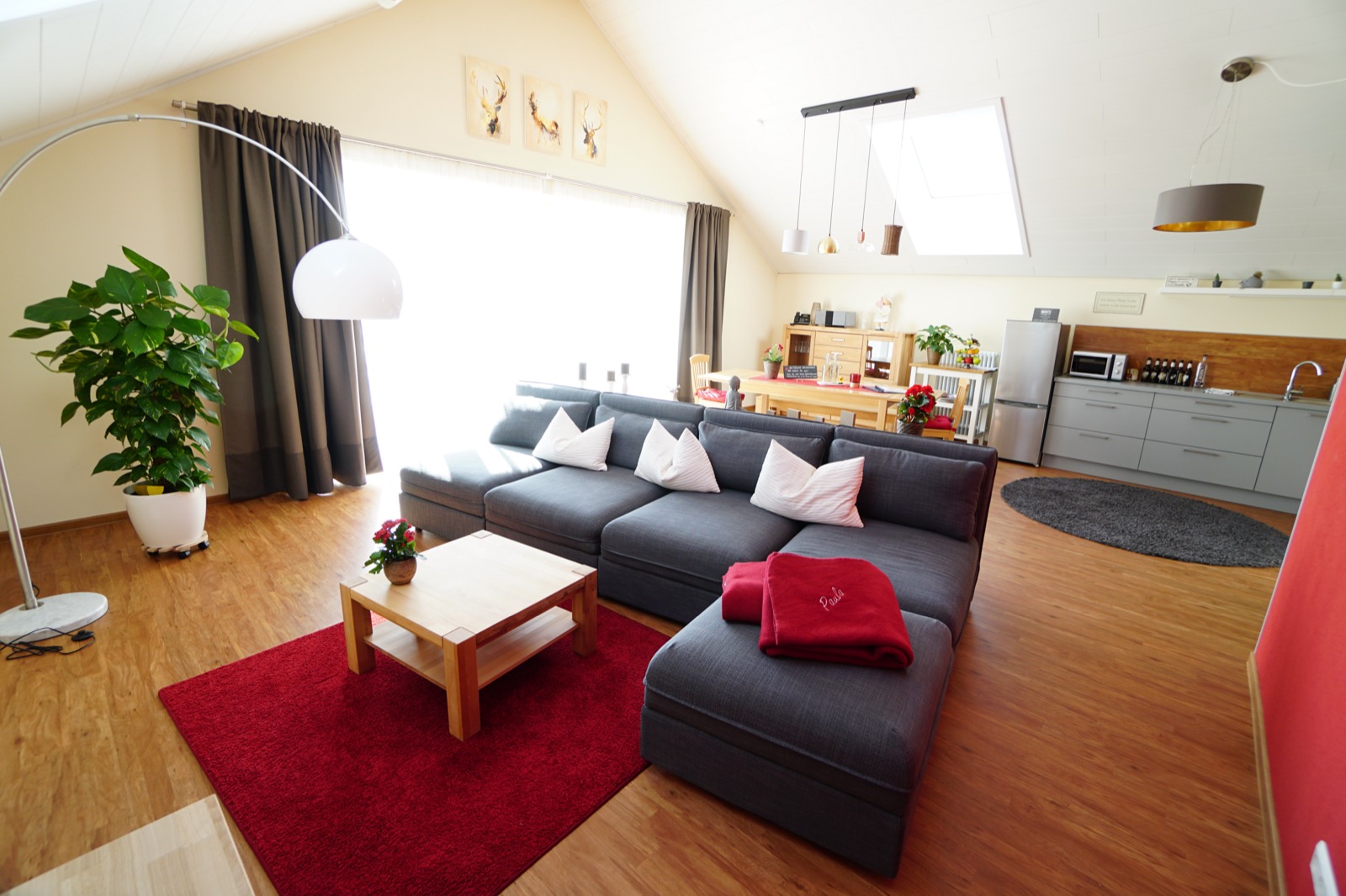 Heller Wohnraum unter der Dachschräge mit großer grauer Couch, rotem Teppich und offener Küche im Hintergrund